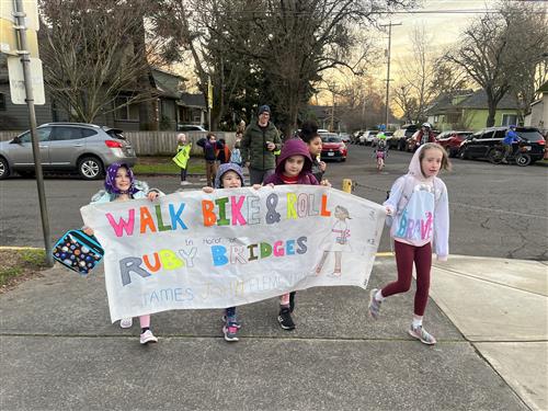 Students walk to school holding a banner reading "Walk Bike & Roll In Honor of Ruby Bridges - James John Elementary"